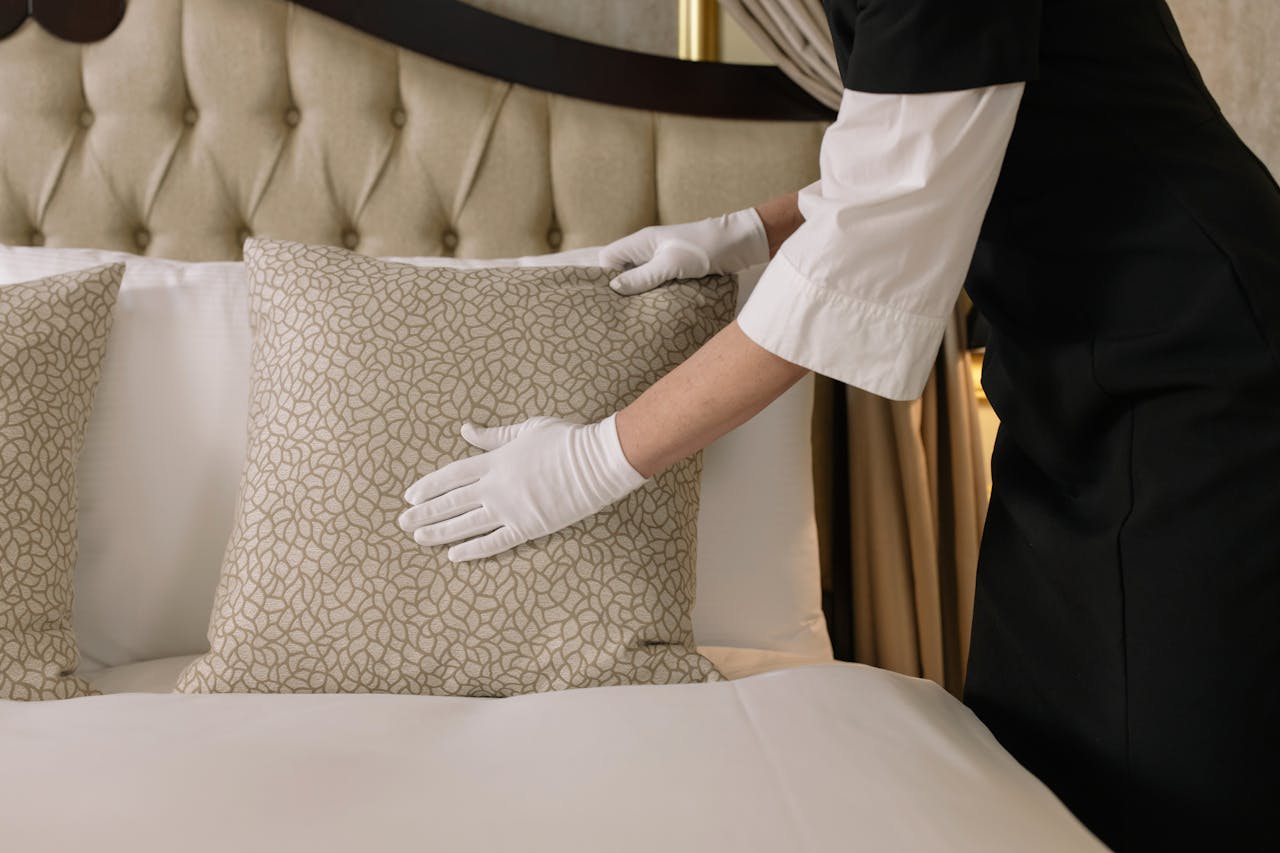 services-04 A housekeeper wearing gloves arranges pillows on a luxurious bed in a hotel room.